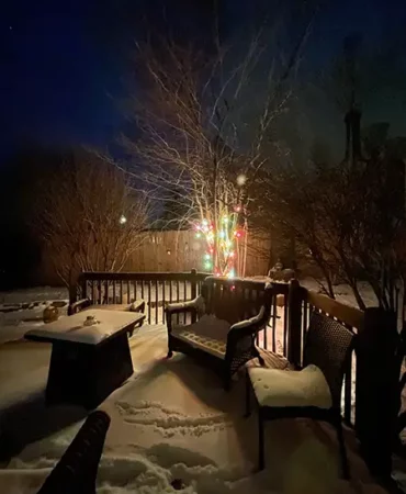Outdoor furniture and decking covered in snow with Christmas lights in the background at night.